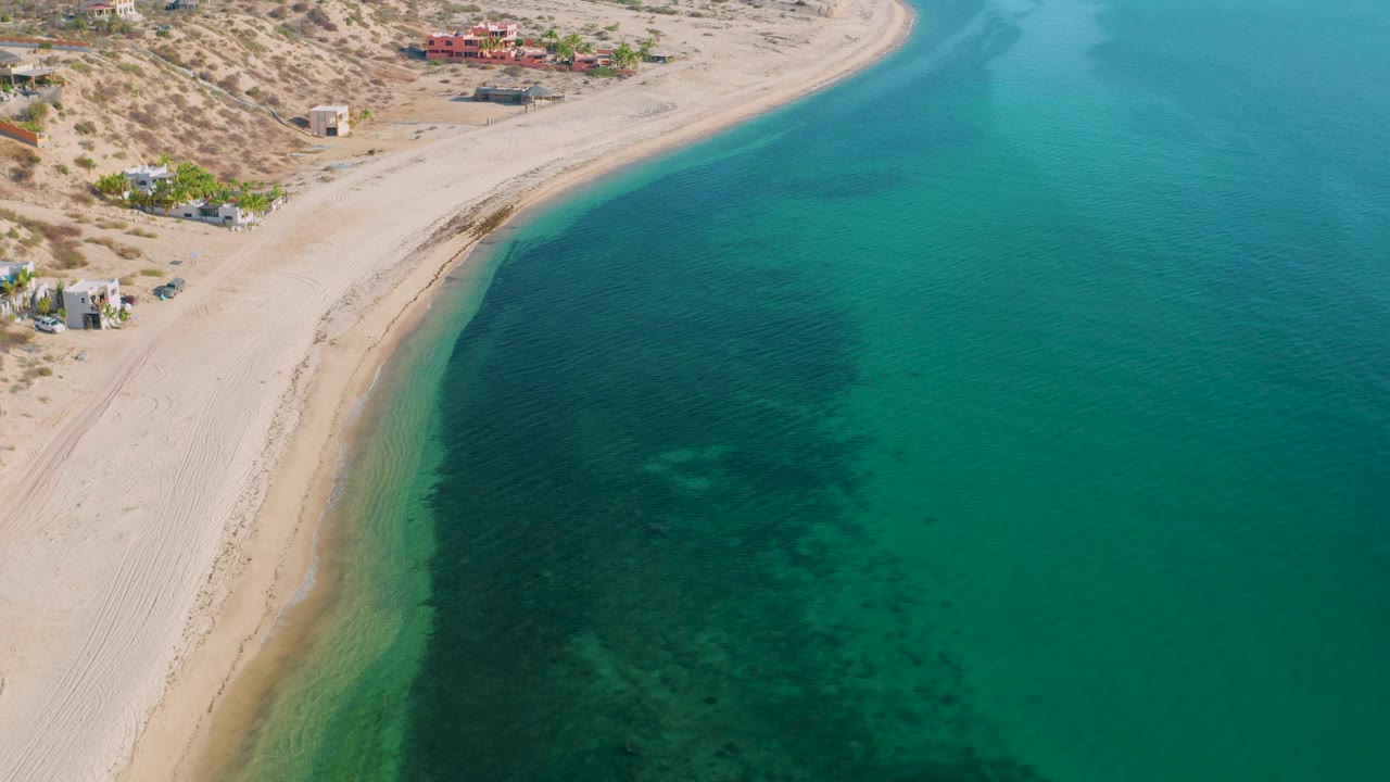 High drone-angle frame in El Sargento La Ventana, Mexico, with the sandy shoreline sweeping upward, turquoise water filling most of the right side, buildings near the upper beach, and no visible people.