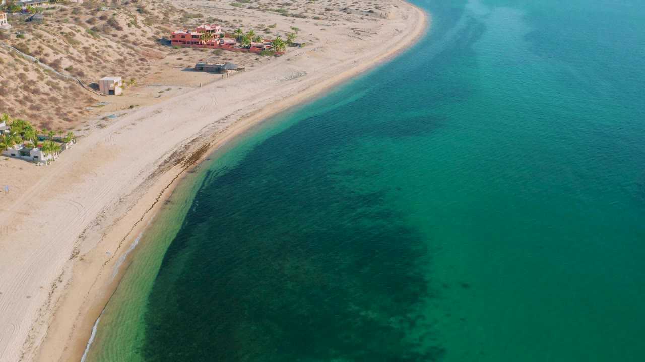 High aerial view in El Sargento La Ventana, Mexico with the beach running diagonally, dark underwater patches near shore, desert brush inland, and no visible people.