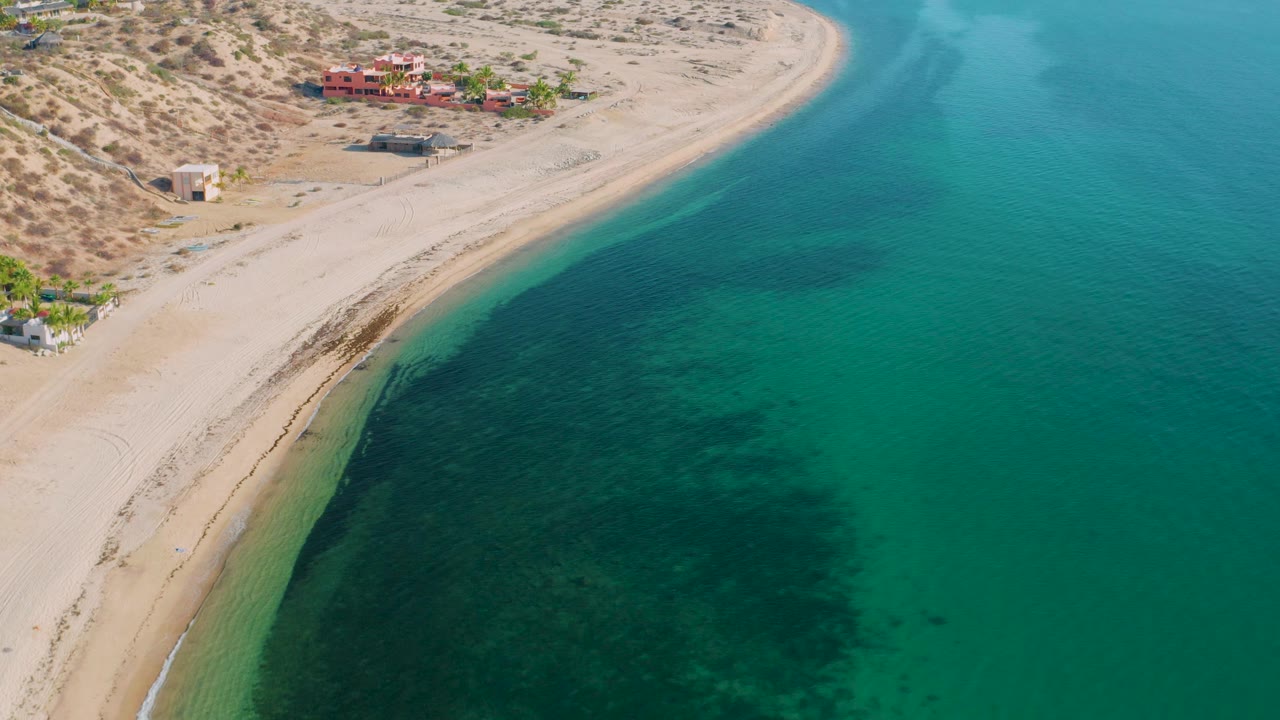 Drone-like overhead frame of El Sargento La Ventana, Mexico with open turquoise water filling the right side, a narrow sandy beach at left, distant buildings, and no visible people.