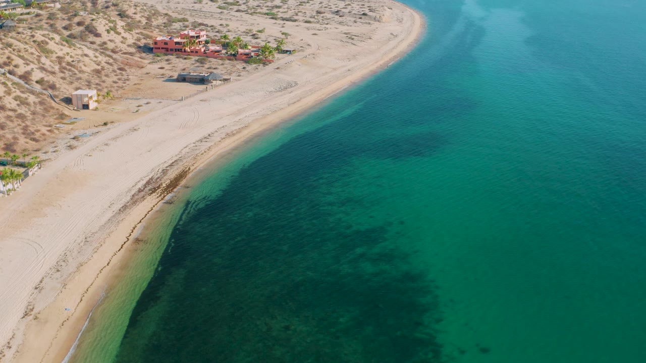 Aerial coastline with pale sand, green-blue water, and low buildings inland.