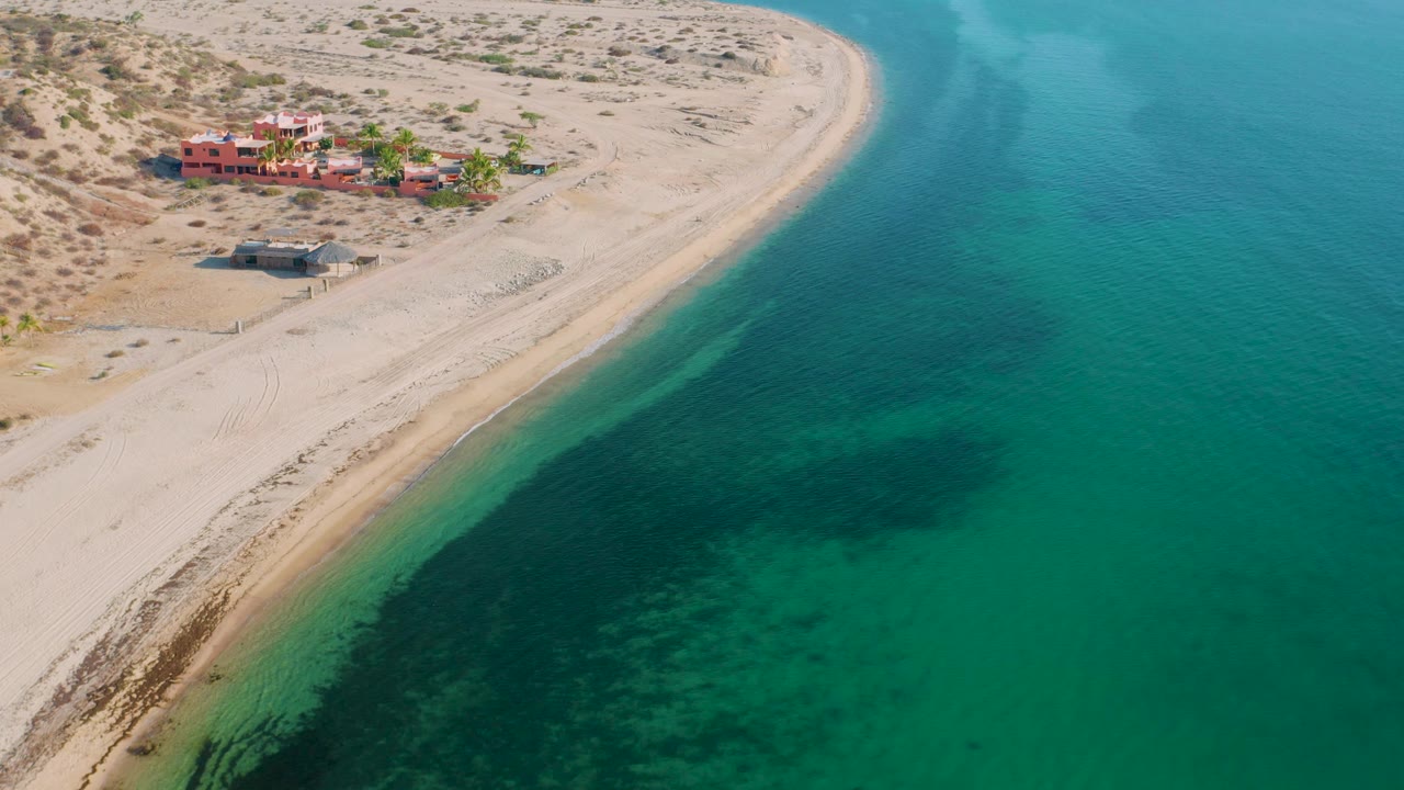 Aerial coastal view in El Sargento La Ventana, Mexico showing beach homes near the upper sand, a narrow pale shoreline, clear turquoise water, darker seabed patches, and no visible people.