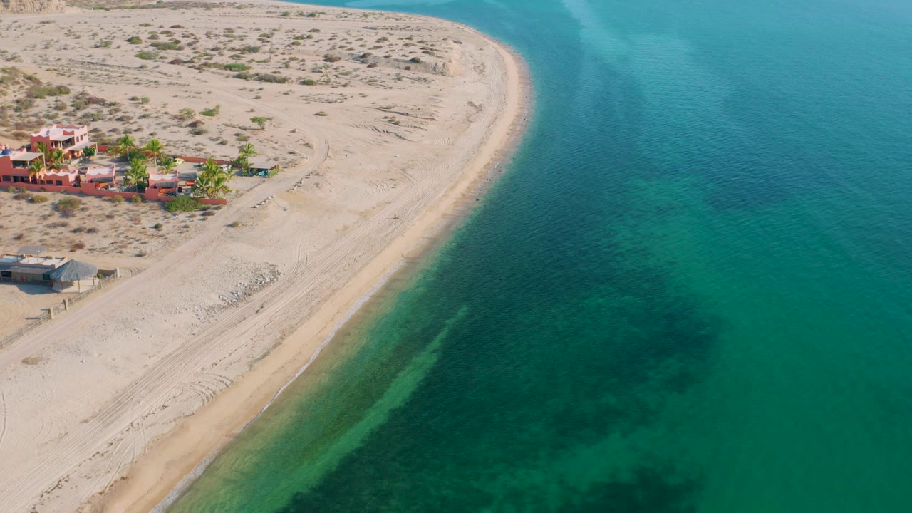 High aerial frame in El Sargento La Ventana, Mexico, with the sandy shoreline cutting diagonally between desert terrain and clear green-blue water, no visible people.