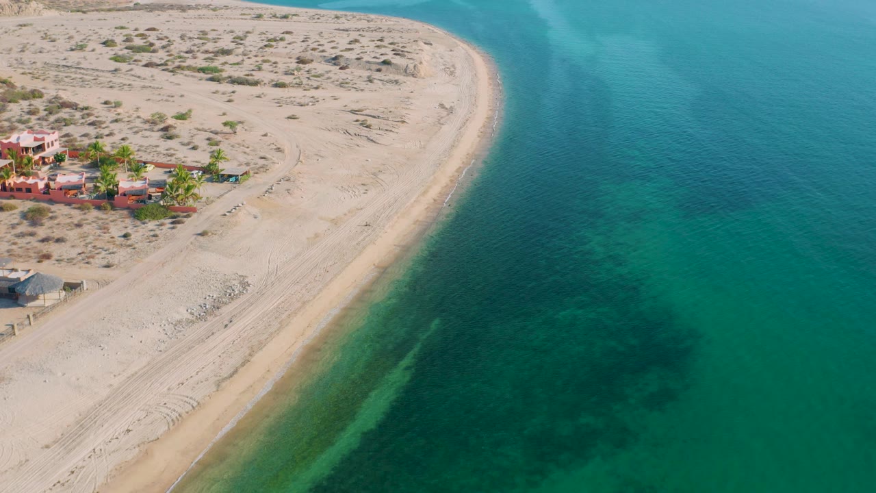 Aerial view over El Sargento La Ventana, Mexico, where a curved sand edge meets green-blue sea with darker underwater areas and beach buildings partly visible at left, no visible people.