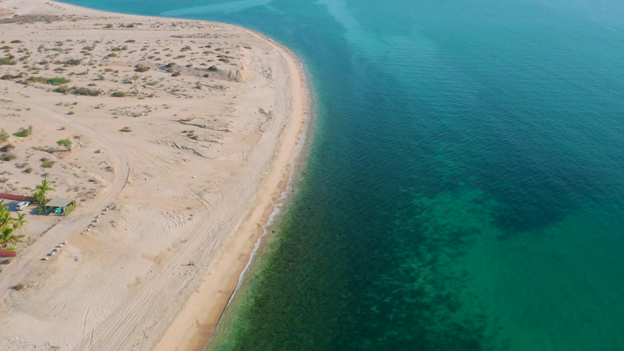Aerial frame over El Sargento La Ventana showing a pale sandy point, sparse scrub, small structures at the far left edge, green ocean water, and no visible people.