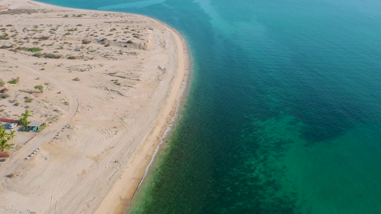 Aerial frame in El Sargento La Ventana with the beach curve centered, pale sand and scrub to the left, darker underwater patches to the right, and no visible people.