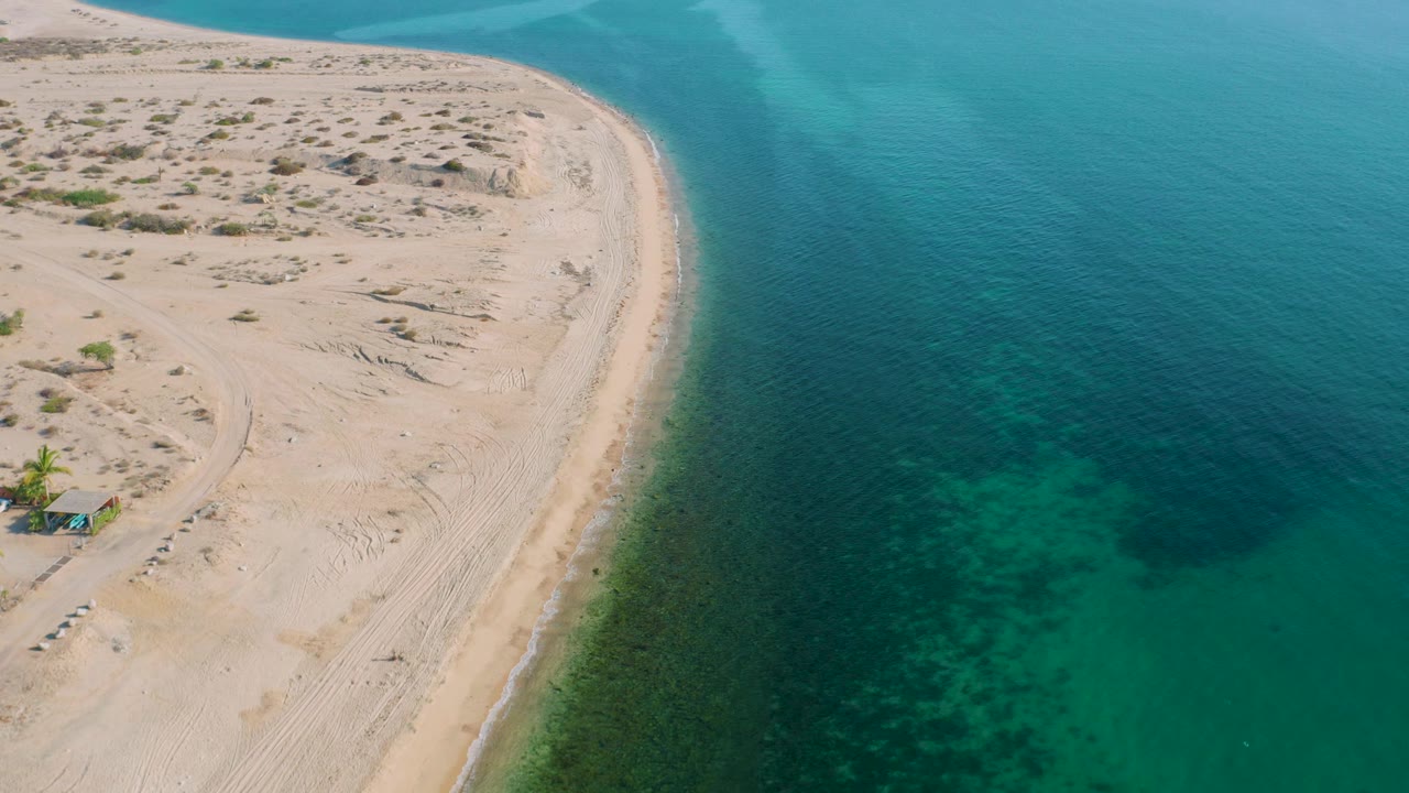 Aerial view over El Sargento La Ventana where the sandy landform narrows toward the top of frame, calm blue-green water fills the right side, and no visible people appear.