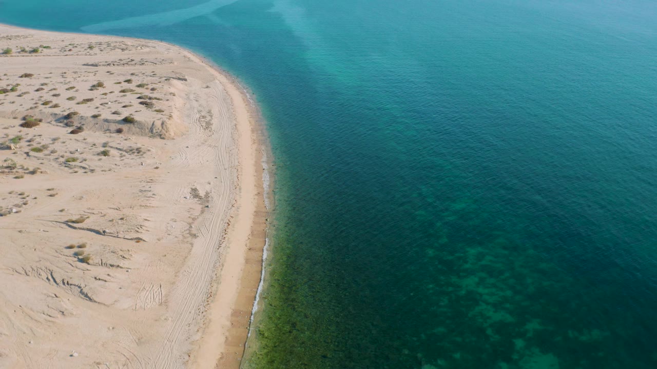 Aerial frame over El Sargento La Ventana, Mexico, with the shoreline slightly farther right and shallow green patches visible under the calm water, no visible people.