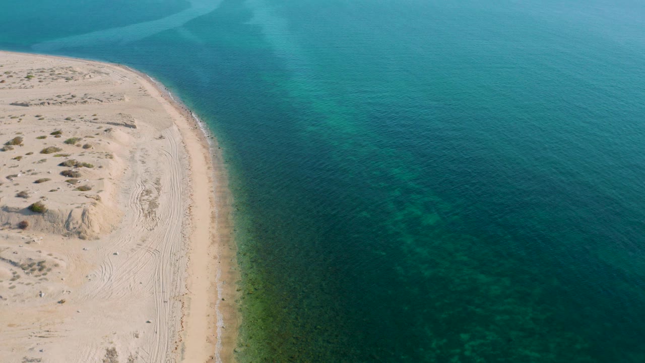 Opening aerial frame of El Sargento La Ventana, Mexico, with a pale sandy point on the left and clear aquamarine water on the right, no visible people.