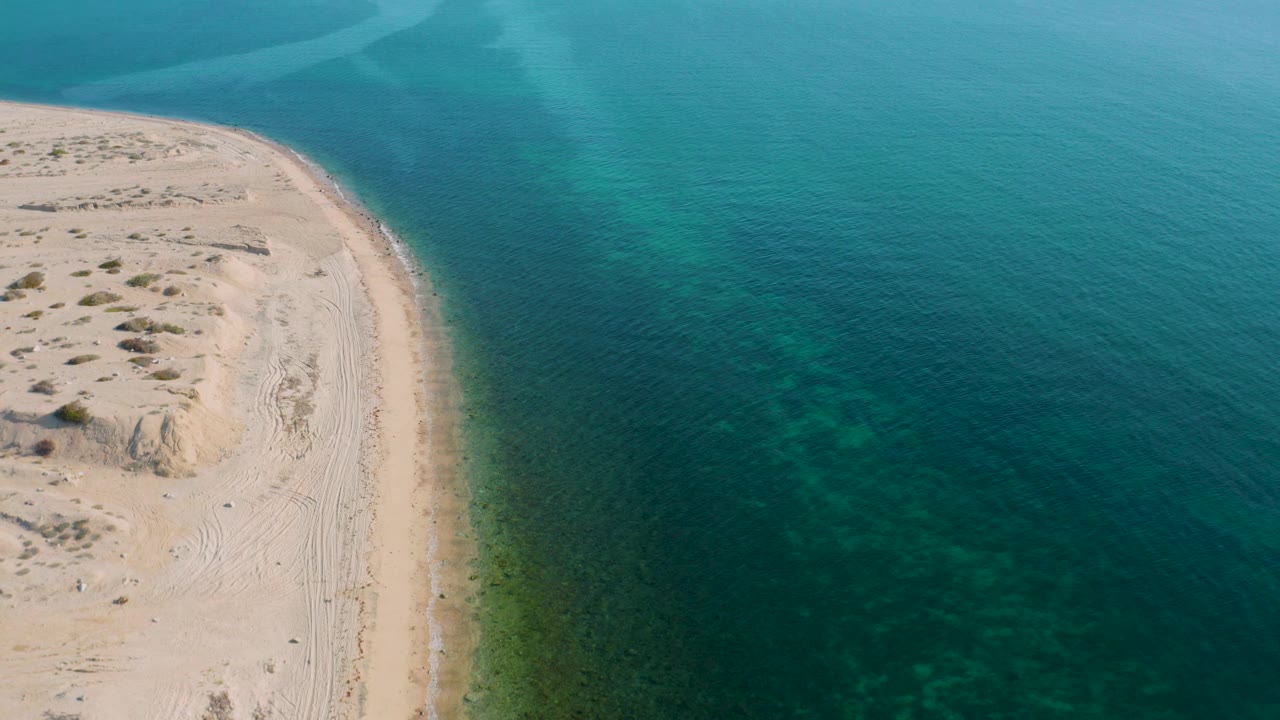 Aerial view over El Sargento La Ventana, Mexico, where the beach arc stays left of frame and dark underwater seaweed patches appear through the blue-green shallows, no visible people.