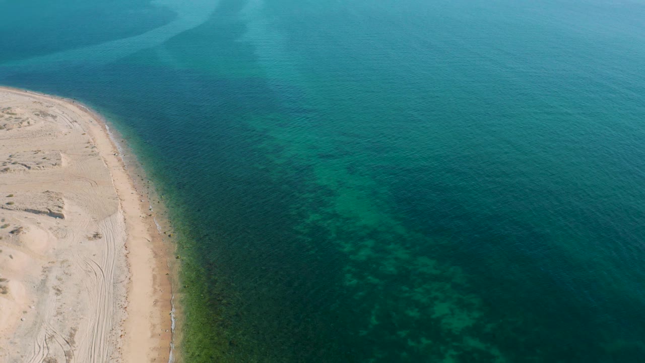 Aerial frame over El Sargento La Ventana, Mexico, showing the curved beach pressed against clear teal water, with a darker band just offshore and no visible people.