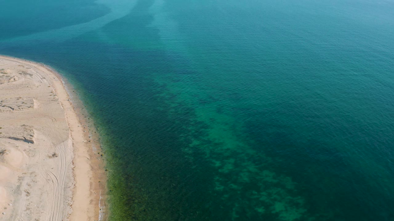 High aerial frame over El Sargento La Ventana, Mexico, with sand occupying the left margin, open teal water across the frame, mottled shallows near shore, and no visible people.