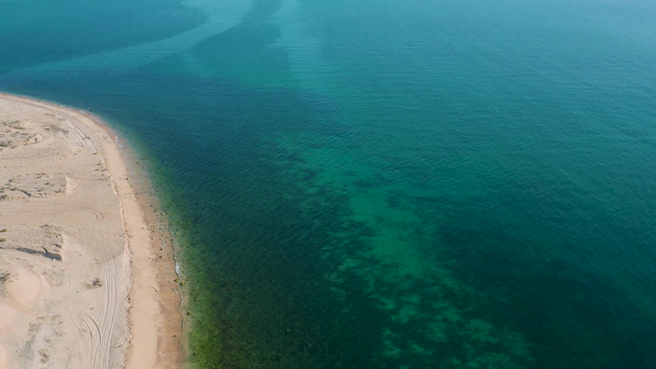 Aerial frame over El Sargento La Ventana, Mexico, where the beach edge sits at left and darker underwater patches spread through the turquoise shallows, with no visible people.