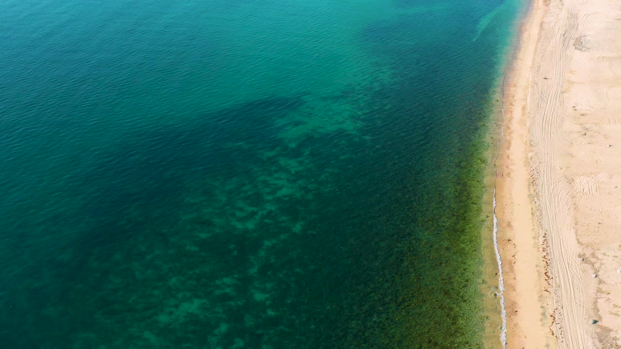 A high aerial frame over El Sargento La Ventana shows blue-green sea, darker underwater patches, and pale sand along the right edge, with no visible people.