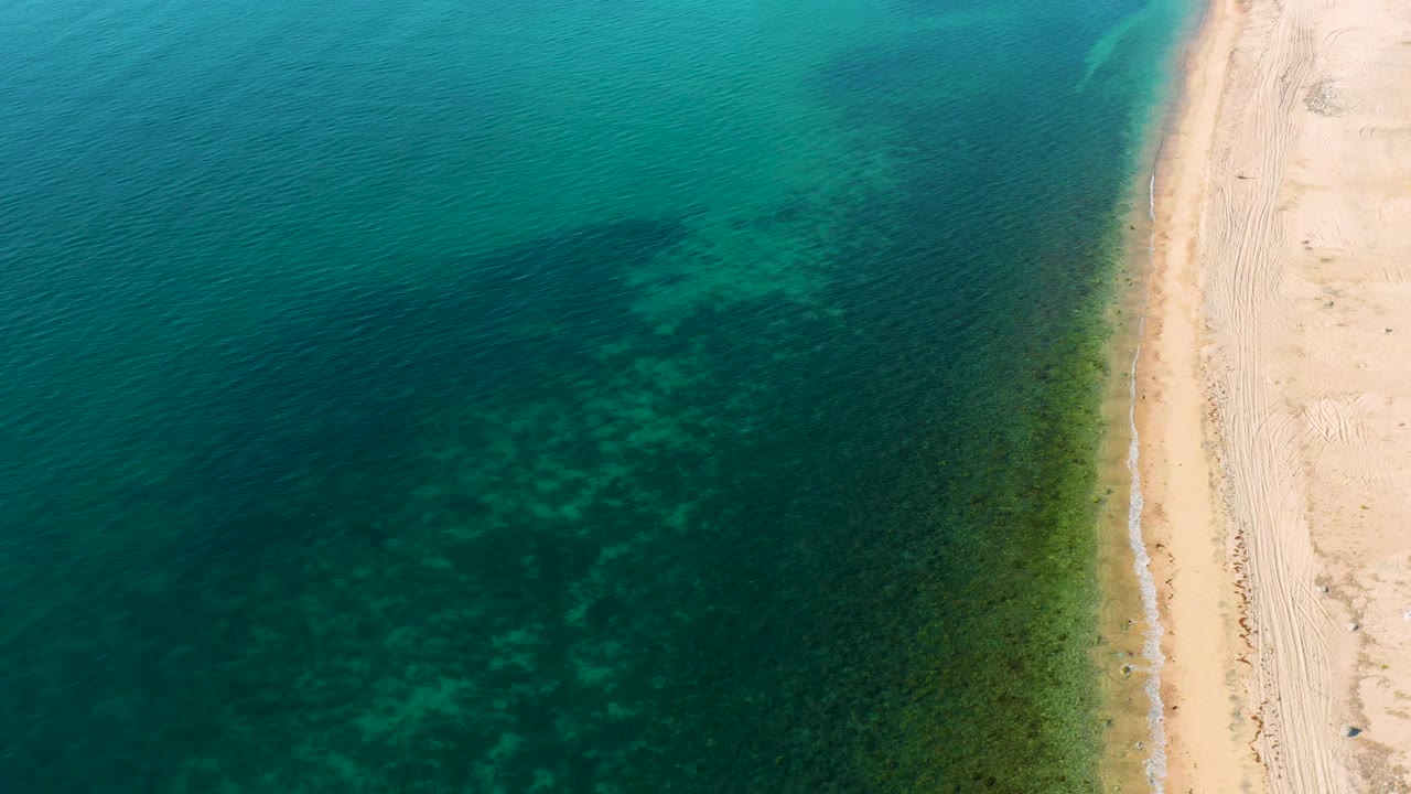 The aerial view over El Sargento La Ventana shows broad blue water, greenish shallows, and a pale sand strip at the right border, with no visible people.