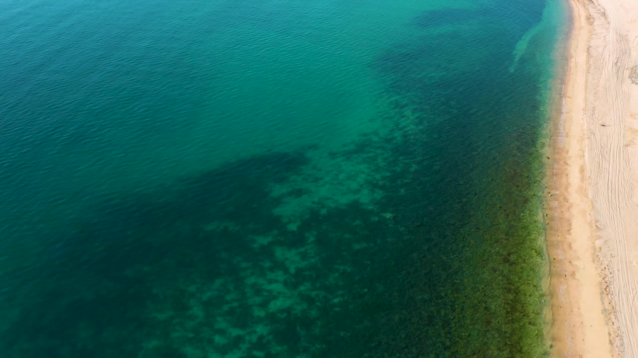 Aerial view over El Sargento La Ventana, Mexico with green-blue shallows, darker reef or seaweed texture beneath the surface, white sand on the right, and no visible people.