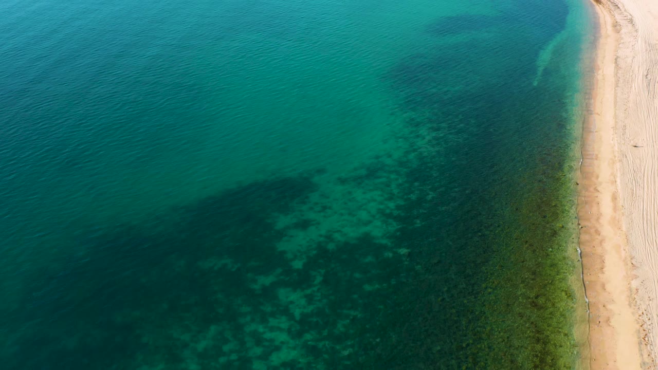 Top-down coastal frame from El Sargento La Ventana, Mexico showing a pale sand strip along the right, translucent turquoise sea to the left, darker underwater areas, and no visible people.