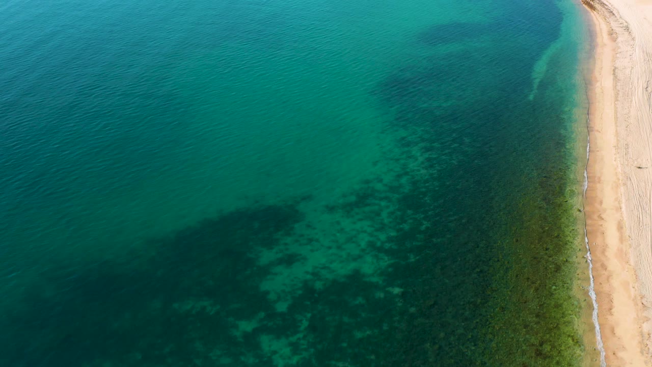 El Sargento La Ventana, Mexico shoreline seen from above, with clear water covering a broad dark underwater patch, a light sand edge on the right, and no visible people.