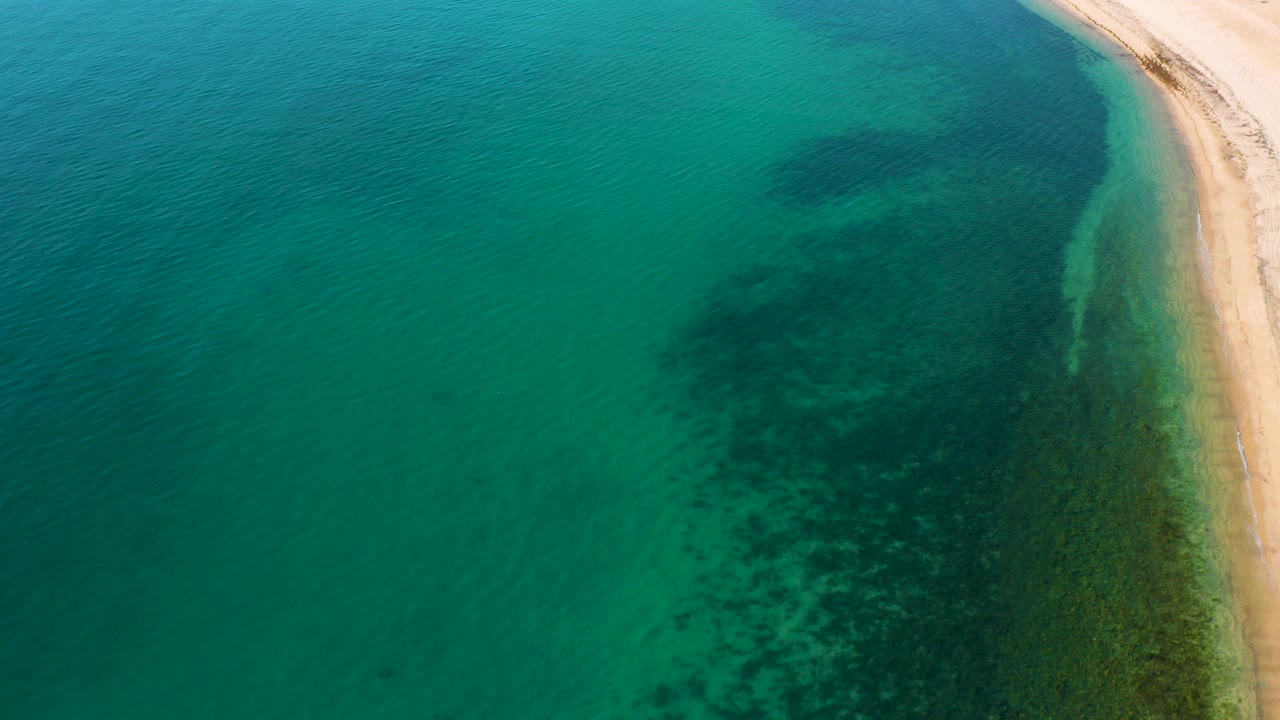 Aerial frame over El Sargento La Ventana, Mexico showing green ocean water, dark underwater patches near the beach, a pale sandy shoreline at right, and no visible people.
