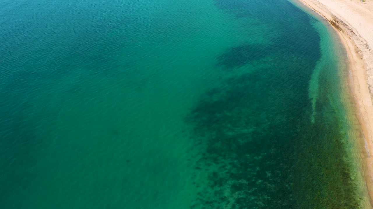 High drone perspective over El Sargento La Ventana, Mexico showing a curved strip of sand at the upper right, clear green water, submerged dark patches, and no visible people.