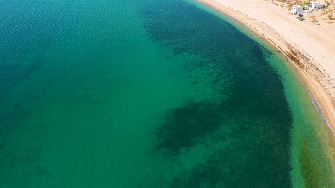 A high coastal view shows clear water over darker shallows beside the pale beach at El Sargento La Ventana, Mexico, with no visible people.