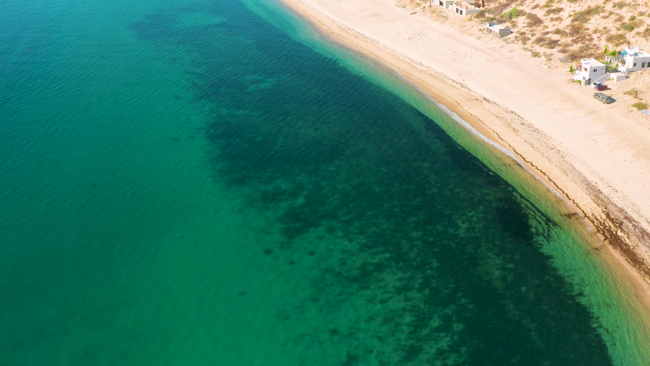 Aerial frame over El Sargento La Ventana, Mexico, showing turquoise shallows, darker underwater patches, a diagonal sand beach, dune scrub, small structures, and no visible people.