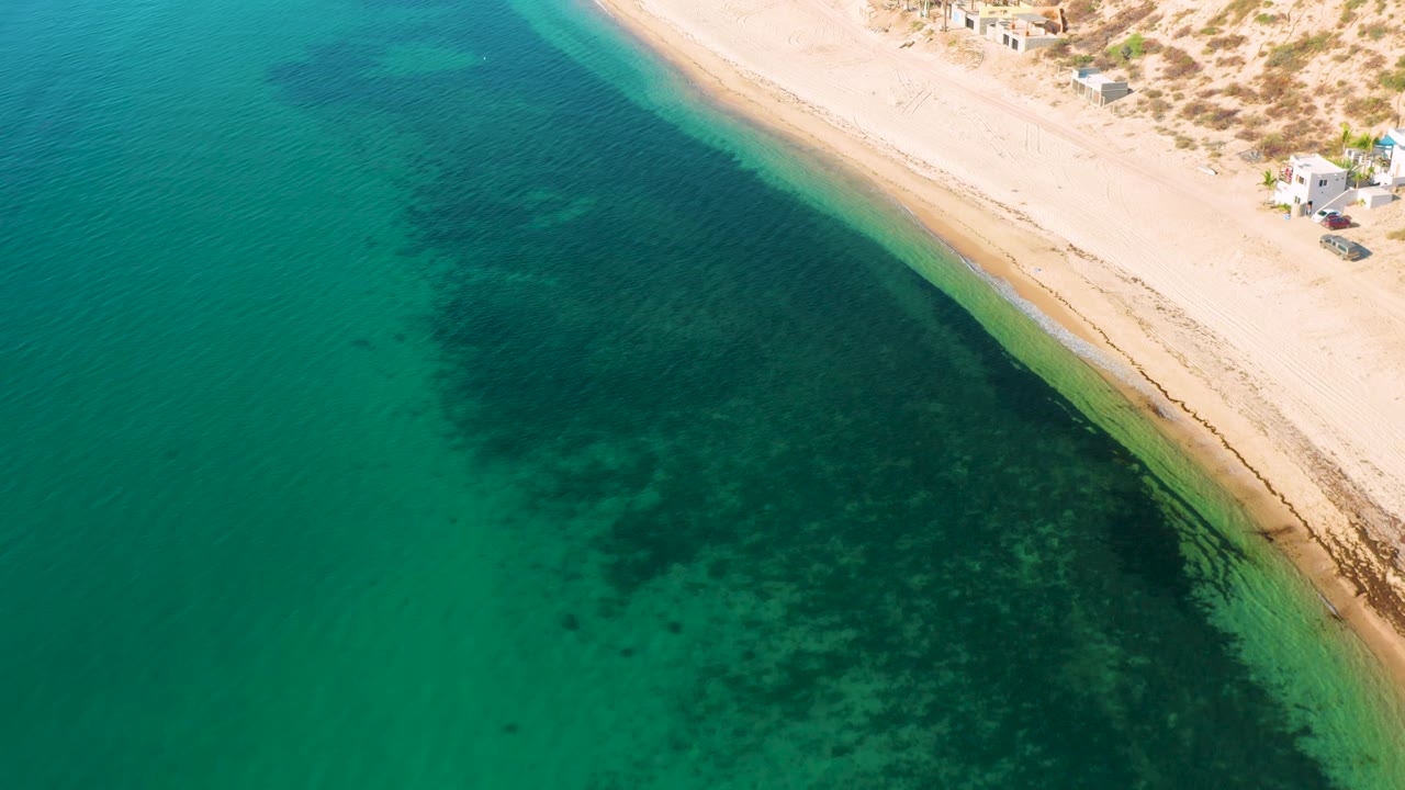 Aerial view of the El Sargento La Ventana shoreline, with a broad dark patch under the clear water, a narrow wet sand edge, dunes and small structures at the top right, and no visible people.