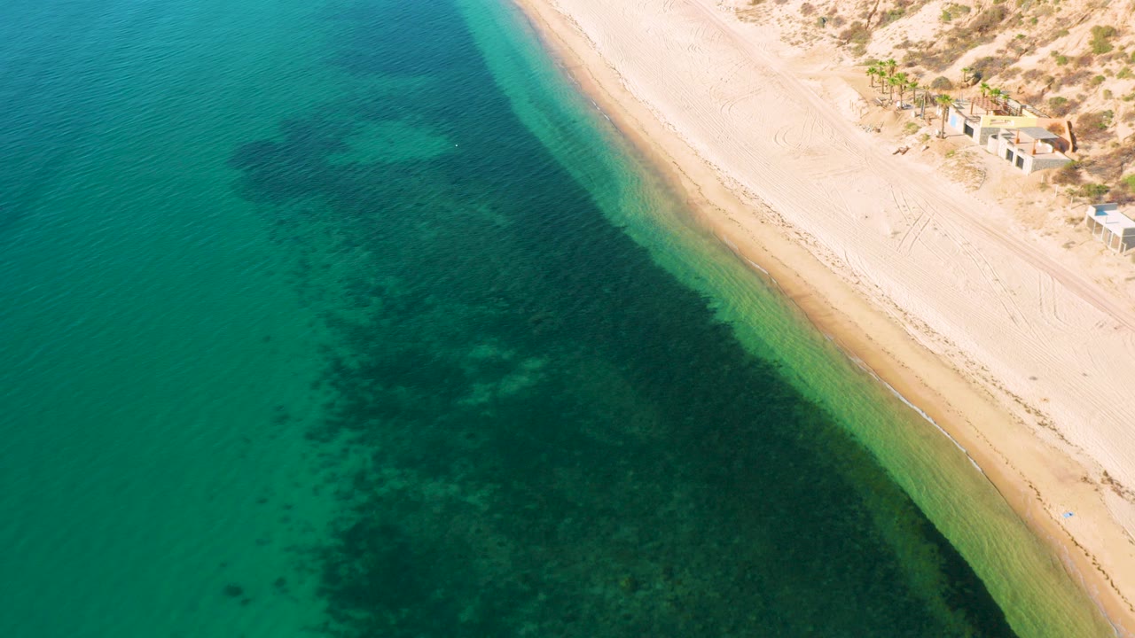 High aerial view of El Sargento La Ventana, Mexico, with turquoise water on the left, pale sand dunes on the right, dark underwater patches near shore, and no visible people.