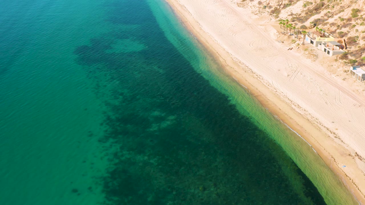 Aerial frame over El Sargento La Ventana, Mexico, showing green-blue water, a narrow pale surf edge, broad sand, dune scrub, and no visible people.