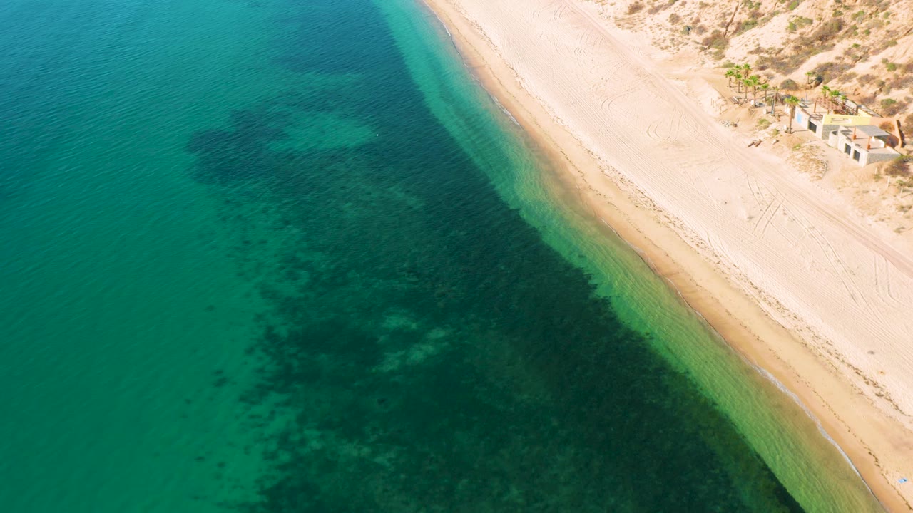 Overhead view in El Sargento La Ventana, Mexico, with clear turquoise ocean, darker underwater formations, a pale sandy beach, dune tracks, small buildings at the upper right, and no visible people.