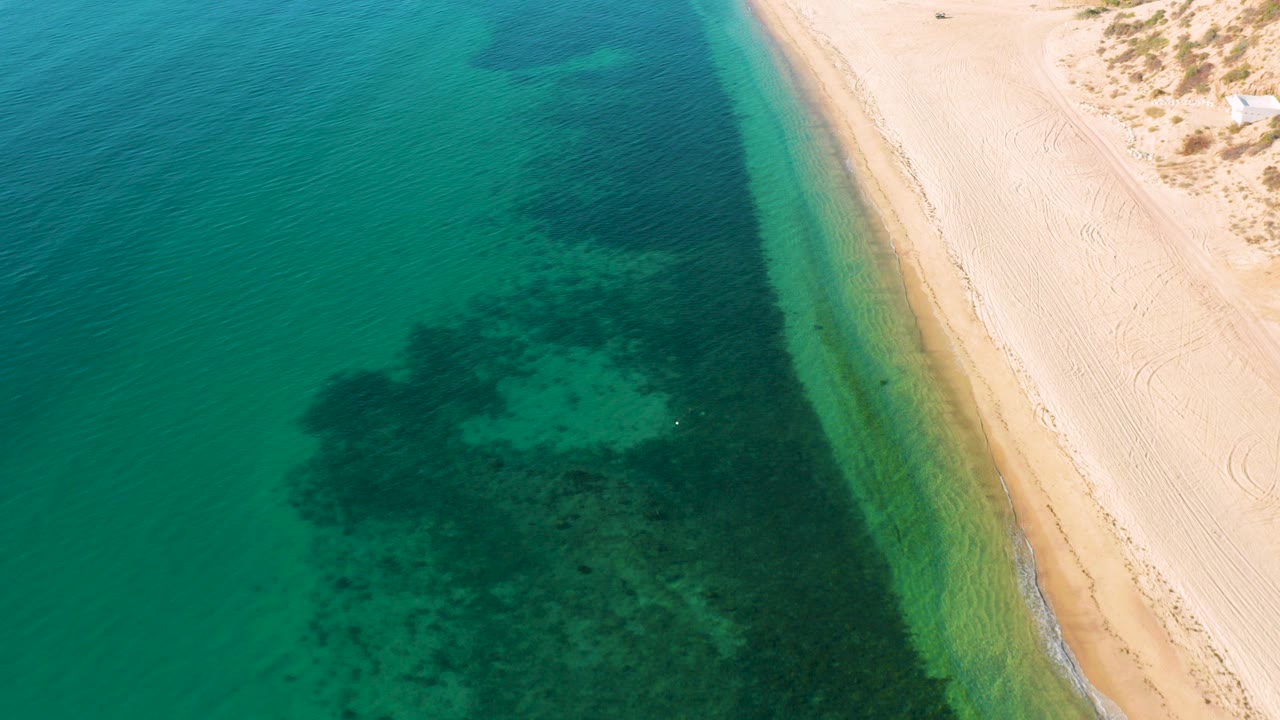 High aerial view over El Sargento La Ventana, Mexico, showing turquoise water meeting pale sand with dark underwater patches and no visible people.