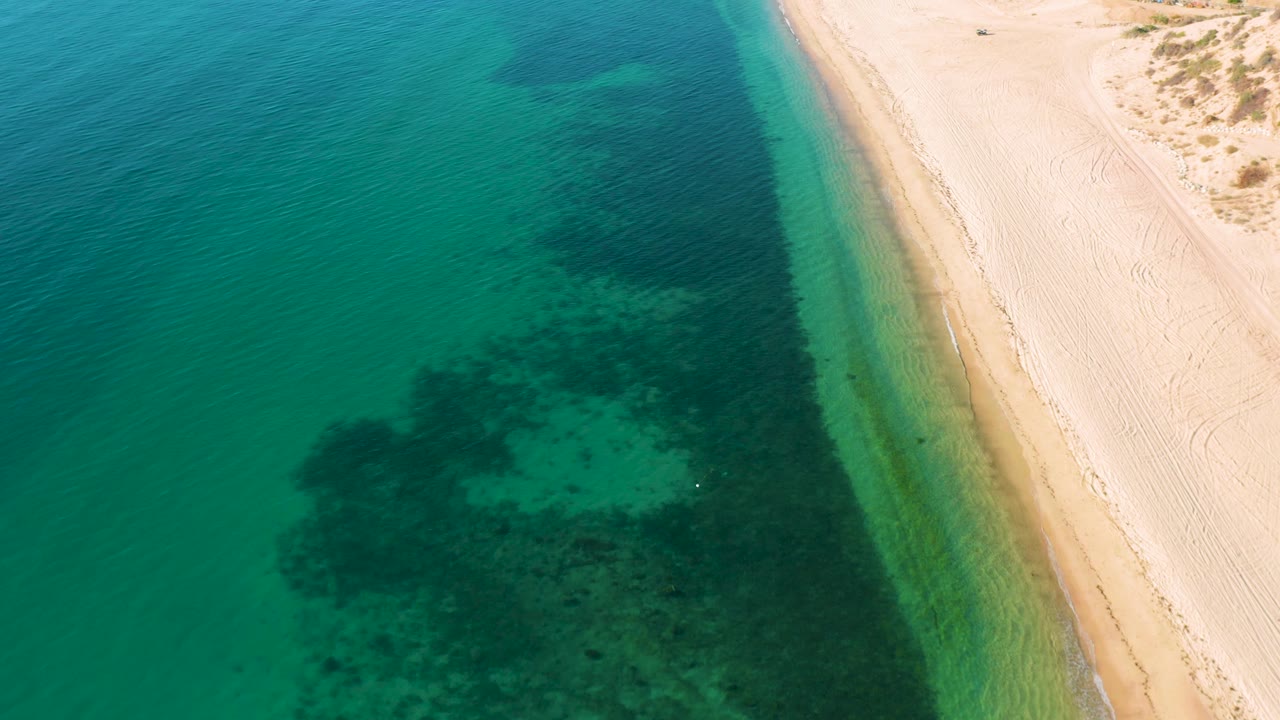 Aerial view over El Sargento La Ventana, Mexico, showing clear shallow water grading into deeper teal beside smooth sand, with no visible people.