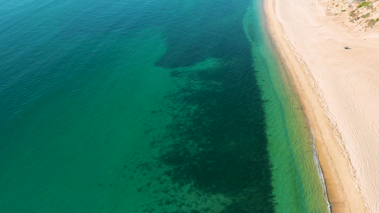 Aerial view of teal water beside pale sand with dark underwater patches