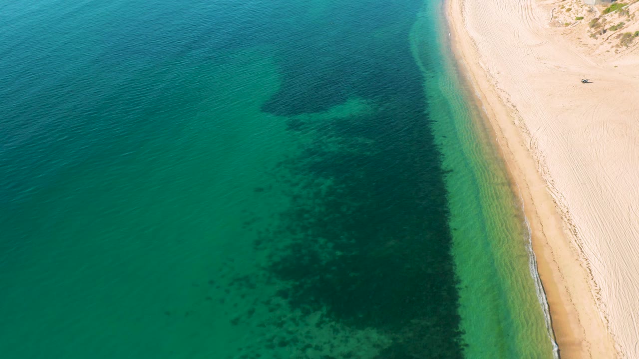 Aerial view in El Sargento La Ventana, Mexico, with a large dark underwater patch just offshore, pale sand along the right edge, and no visible people.