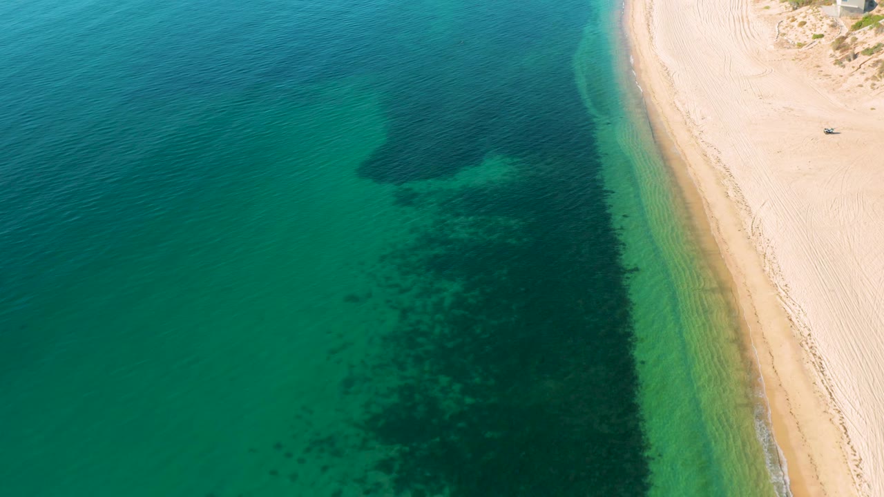 High drone-style frame over El Sargento La Ventana, Mexico, showing green-blue water meeting a diagonal strip of pale sand, with no visible people.