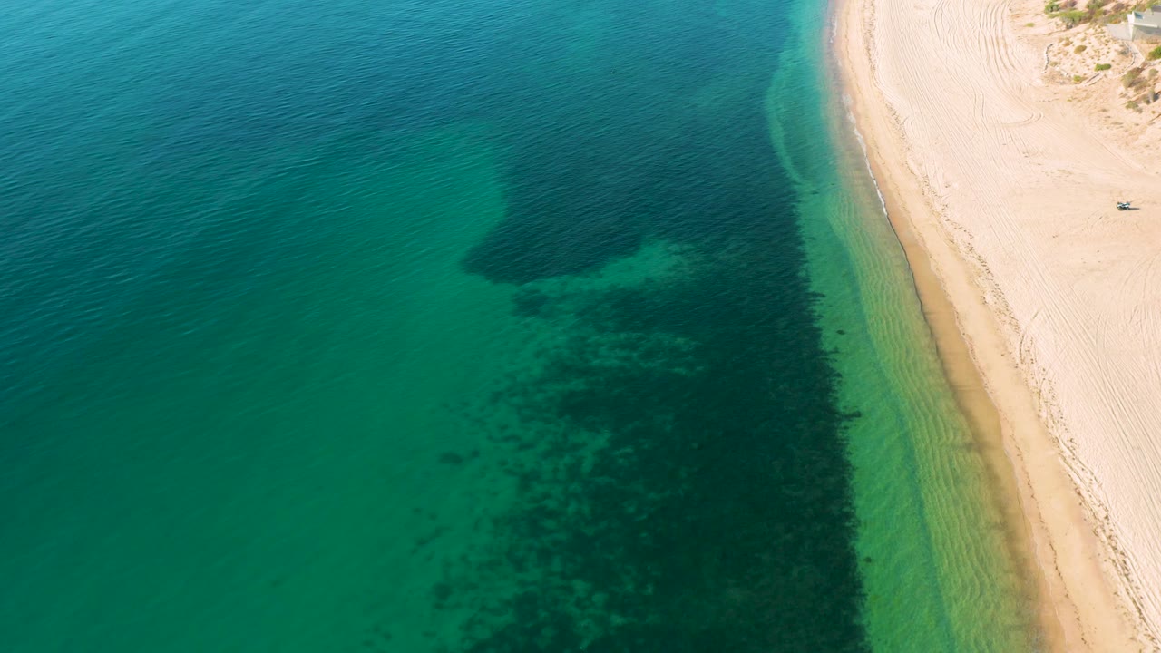 Aerial frame from El Sargento La Ventana, Mexico, with clear coastal water, a rounded dark seabed shape near the beach, pale sand, and no visible people.