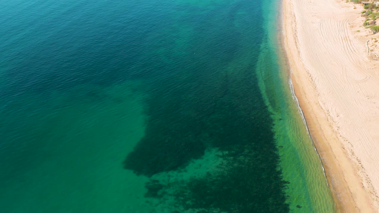 Overhead frame from El Sargento La Ventana, Mexico showing teal water on the left, a darker submerged patch near the lower center, pale sand on the right, and no visible people.