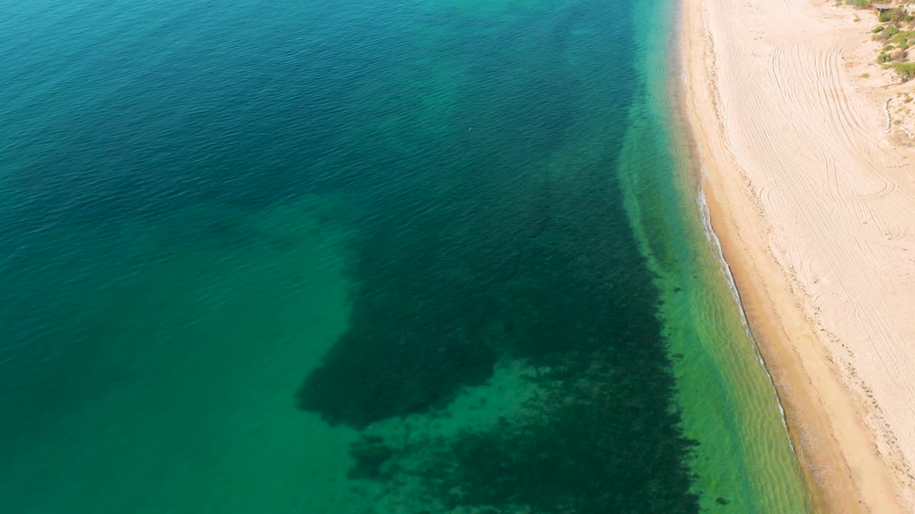 Aerial frame from El Sargento La Ventana, Mexico with dark green underwater texture expanding near the lower middle, shallow water along the beach, pale sand at right, and no visible people.