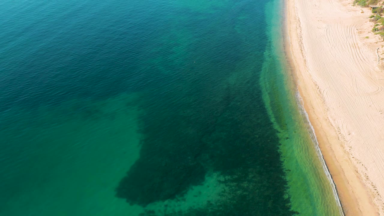 High overhead view from El Sargento La Ventana, Mexico showing a larger field of clear green water, darker seabed shapes, a thin wave line, pale sand, and no visible people.