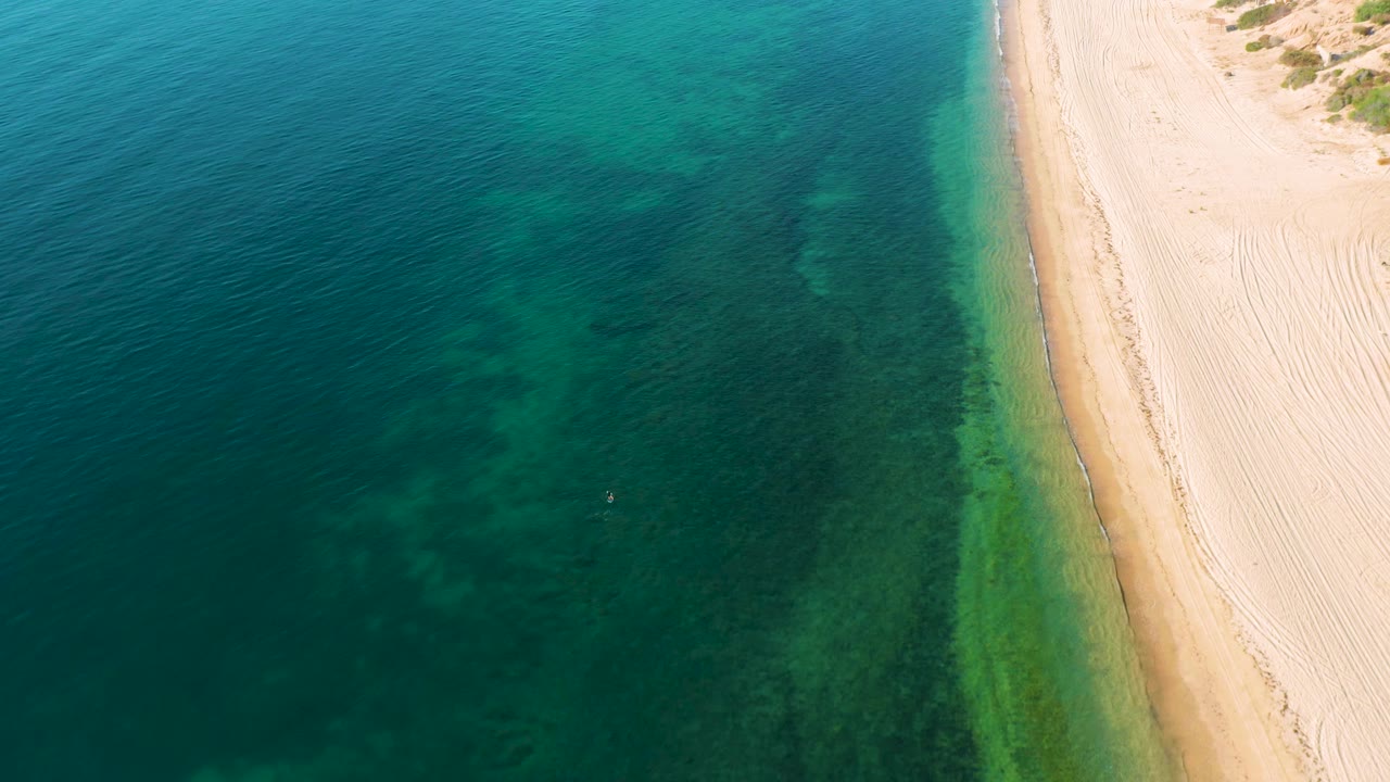 Aerial frame over El Sargento La Ventana, Mexico, showing blue-green water with submerged darker reef or sea grass texture and a bright sand strip on the right, no visible people.