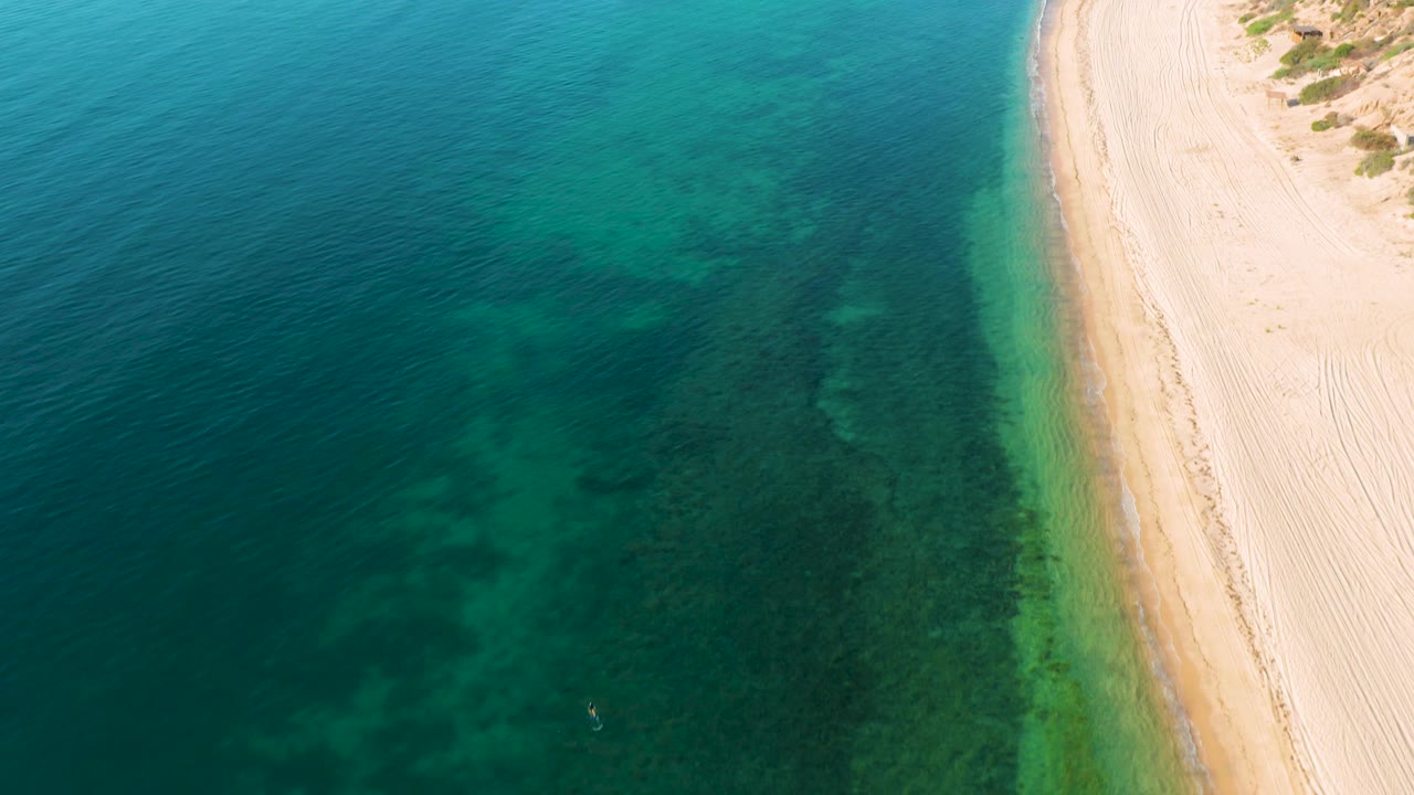 Aerial shoreline view from El Sargento La Ventana, Mexico, with transparent water darkening toward the left and a slim pale beach edge on the right, no visible people.
