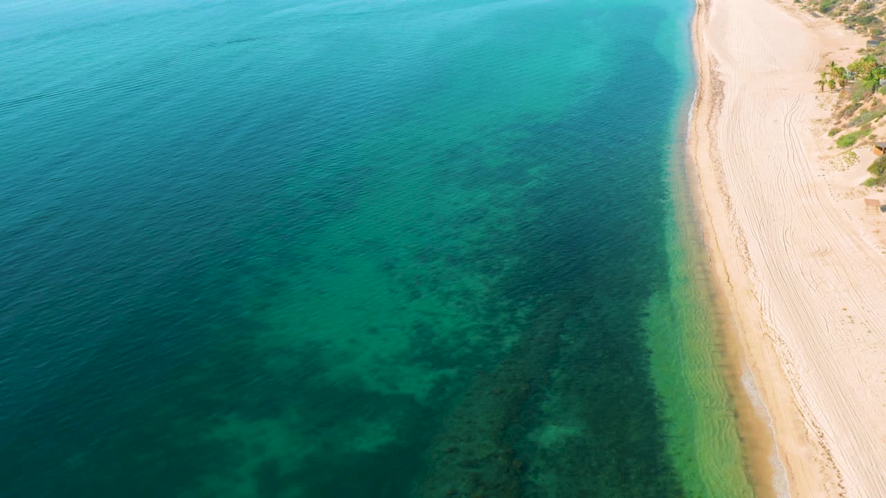 Aerial frame of blue-green water and a narrow sandy shoreline in El Sargento La Ventana, Mexico, with the beach held tight to the right side and no visible people.