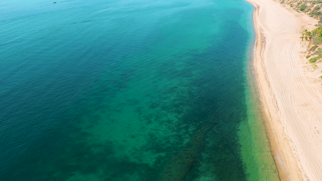 High view over clear coastal water in El Sargento La Ventana, Mexico, with pale sand on the right, hints of shoreline vegetation near the top, and no visible people.