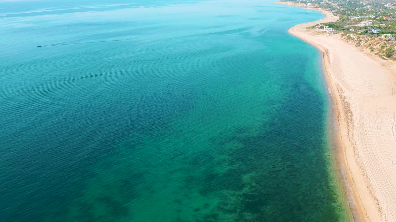 Aerial view in El Sargento La Ventana, Mexico with blue-green water across most of the frame, a pale curving shoreline on the right, darker underwater patches near the lower center, and no visible people.