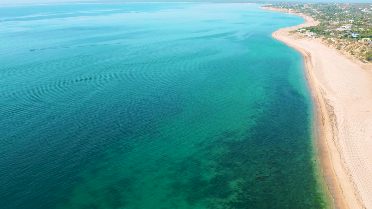High aerial frame over El Sargento La Ventana, Mexico showing turquoise sea with darker submerged reef shapes, pale sand along the right edge, scattered shoreline buildings beyond it, and no visible people.