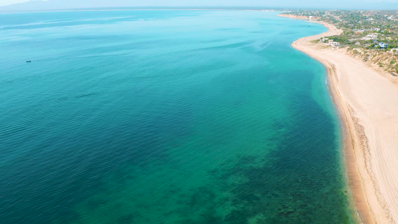 Aerial frame in El Sargento La Ventana, Mexico with fine ripples on turquoise water, darker reef areas below, a narrow pale beach at right, distant coast at top, and no visible people.