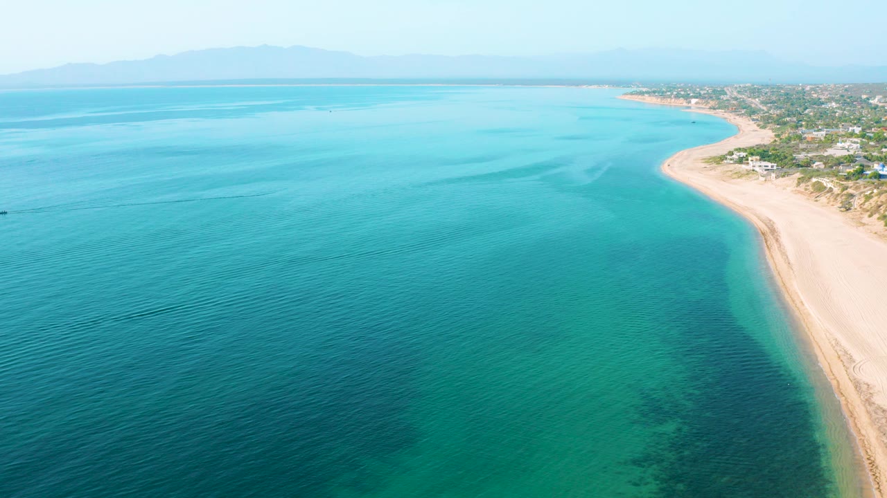 High aerial view over turquoise water with a pale beach curving along El Sargento La Ventana, Mexico, coastal homes and trees at right, hazy mountains on the horizon, and no visible people.