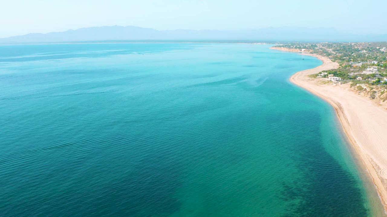 Wide overhead coastal view in El Sargento La Ventana, Mexico, showing blue-green water across the frame, a curved sandy beach at right, shoreline vegetation, small buildings, distant mountains, and no visible people.