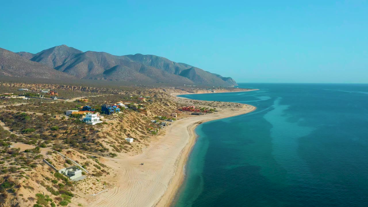 A high aerial view over El Sargento La Ventana, Mexico shows scrubby dunes and scattered coastal buildings on the left, a pale beach curving beside turquoise water on the right, mountains in the distance, and no visible people.