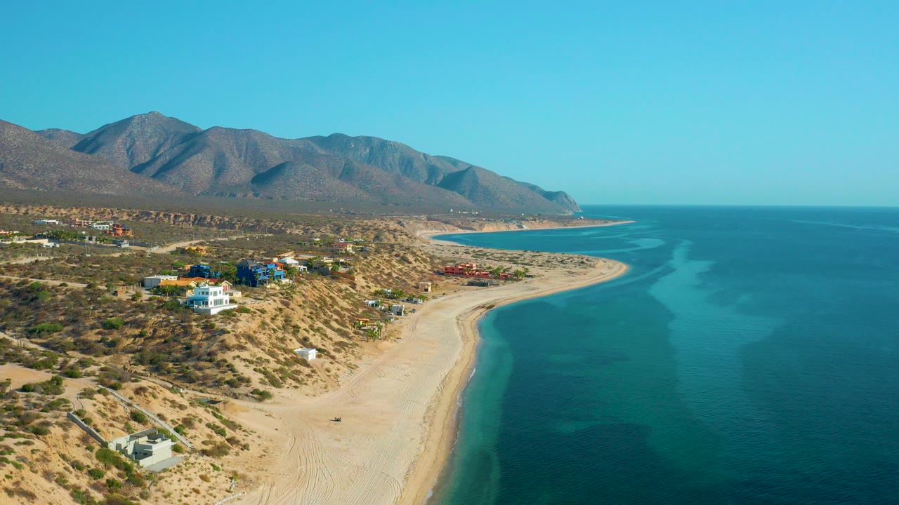 Opening aerial frame over El Sargento La Ventana, Mexico, with pale sand curving along blue water, scattered homes on scrubland, mountains beyond, and no visible people.