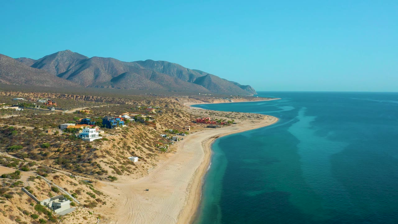 The frame shows El Sargento La Ventana, Mexico from above, with the sandy beach and bright water filling the right side and small coastal buildings set into dry terrain at left, with no visible people.